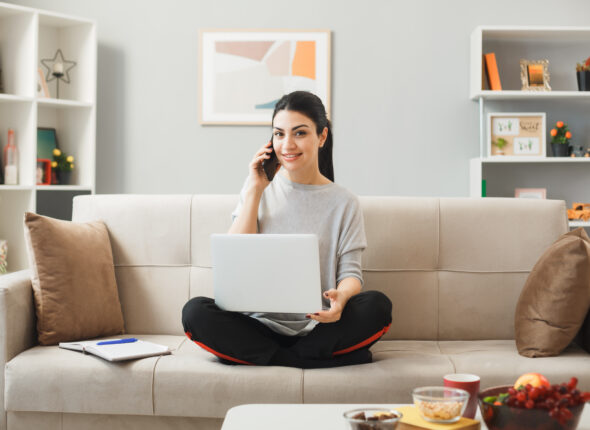 young-woman-holding-used-laptop-speaks-phone-sitting-sofa-coffee-table-living-room